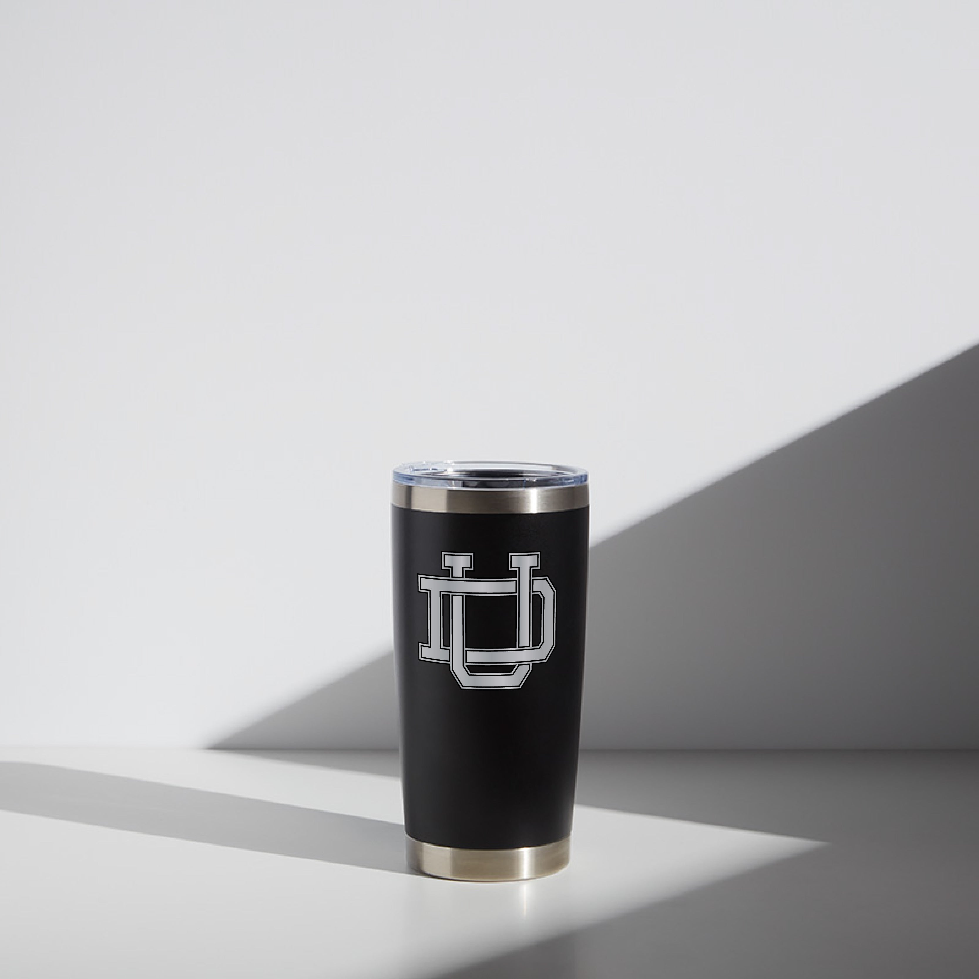 Alt text: Black 20 oz WATR Tumbler featuring the Dayton Interlocking UD logo in white, set against a simple backdrop with a prominent shadow, showcasing the metallic rim and clear lid.