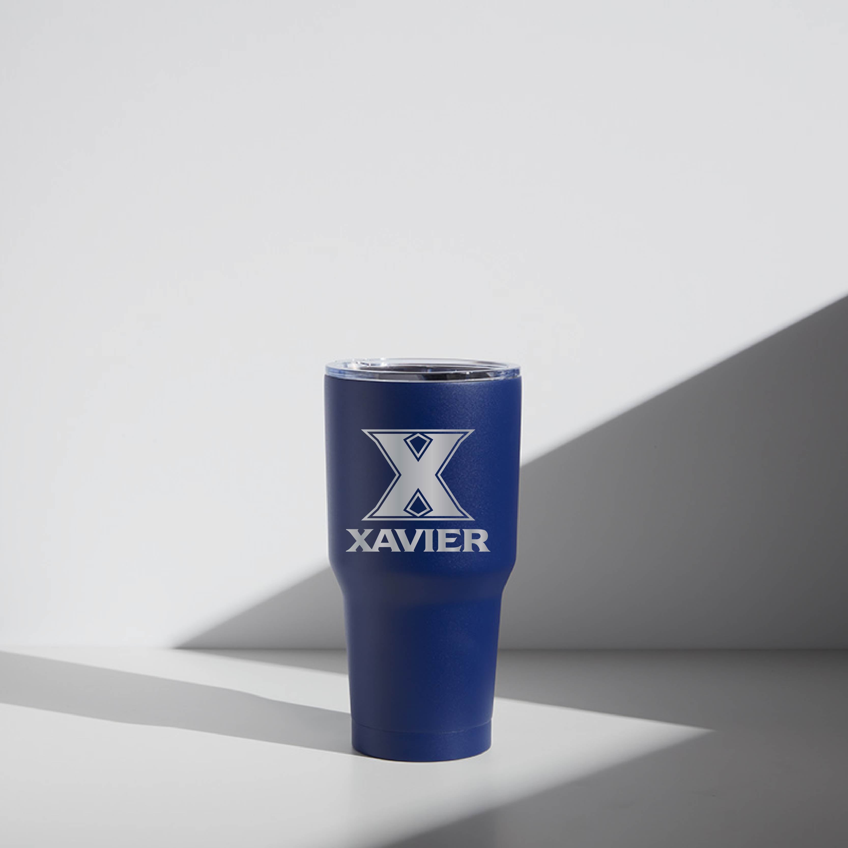A blue 30 oz stainless steel tumbler with the text 'XAVIER' printed on it, placed against a white background with a shadow cast beneath it.