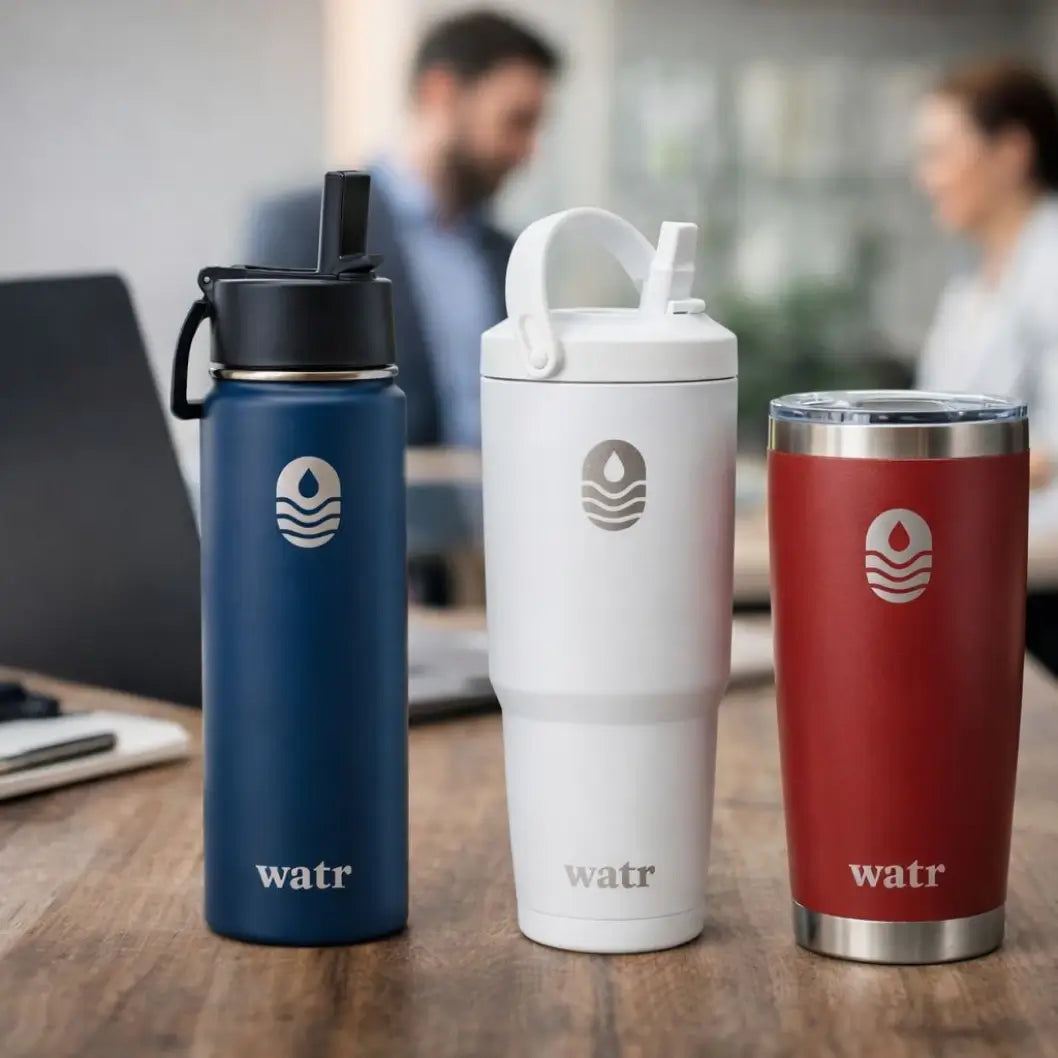 Three WATR insulated drinkware products displayed on a wooden conference table — a navy water bottle, a white tumbler with a handle, and a red tumbler — with two people meeting in the background